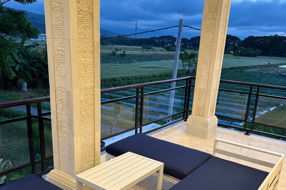 A balcony with a view of a green landscape, a white table, and blue cushioned seating.