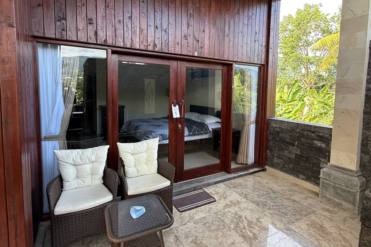 A room with a wooden ceiling, a glass door leading to a bedroom, a wicker sofa with cushions, and a tiled floor.
