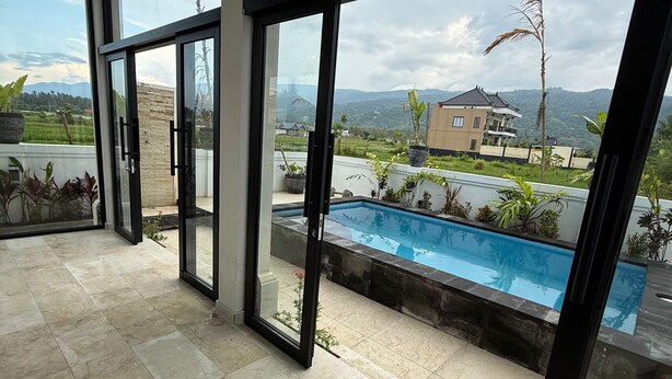 A modern interior with large glass doors leading to a pool area, a tiled floor, and a view of a residential area with a house and mountains. - Murni's House (Buleleng)