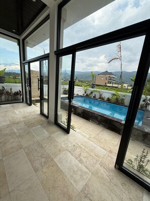 Un interior moderno con grandes puertas de cristal que dan a una zona de piscina, suelo de baldosas y vistas a un área residencial con una casa y montañas.