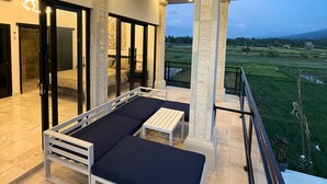 A modern outdoor seating area with a sofa and table, a glass door entrance, and a view of a green landscape. - Murni's House (Buleleng)