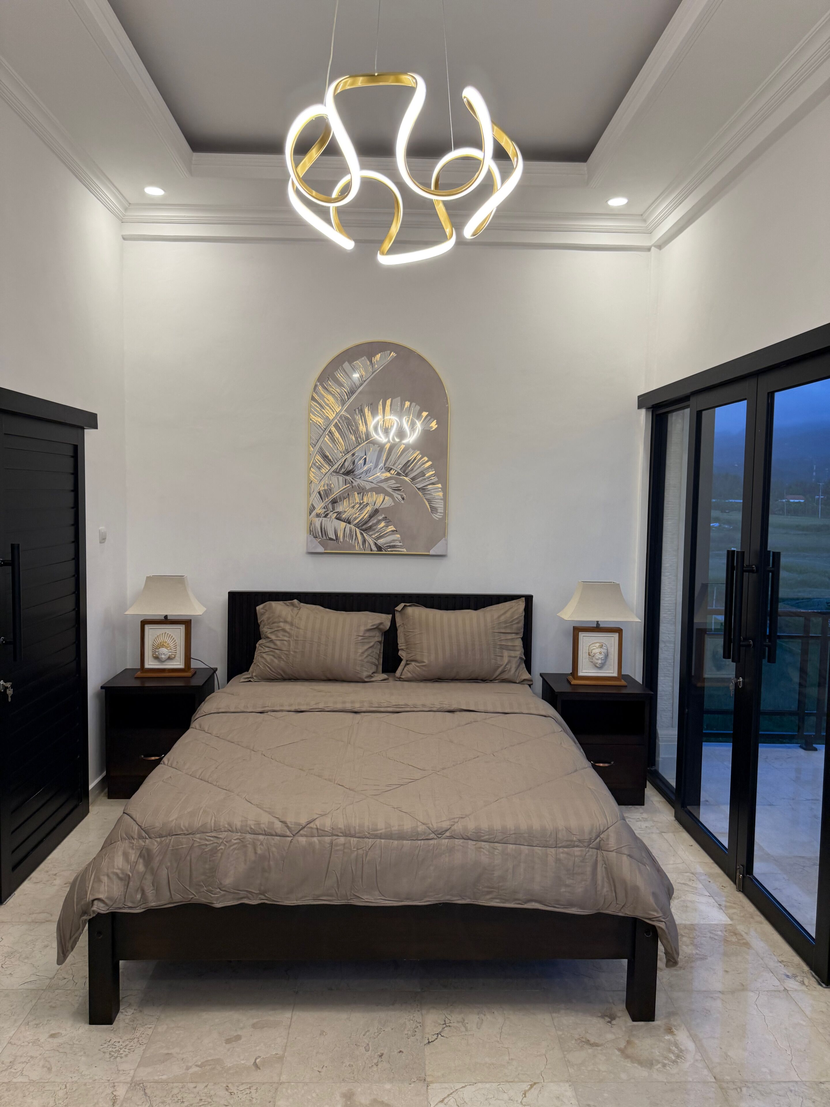 A modern bedroom with a large bed, two bedside tables with lamps, a wall-mounted artwork, and a sliding glass door to a balcony.