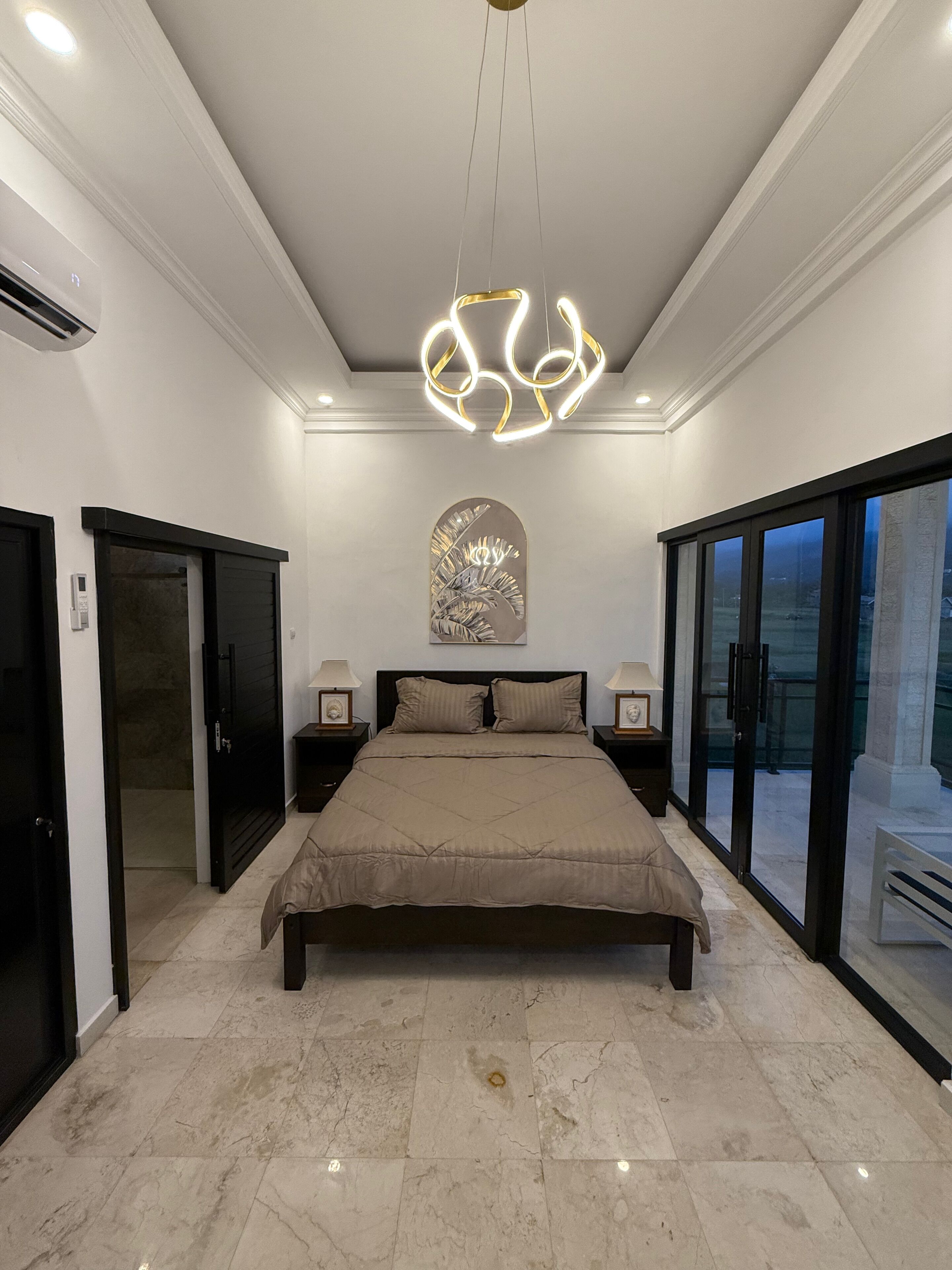 Un dormitorio con una cama grande, un candelabro en el techo, una obra de arte en la pared y un ventanal corredero que da a un balcón.