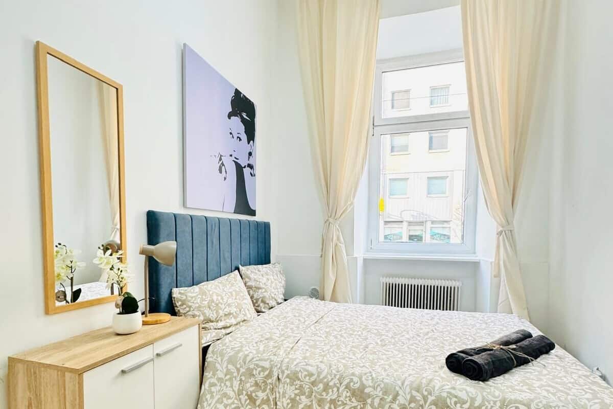 Een slaapkamer met een bed, nachtkastje, spiegel, raam met gordijnen en een kunstwerk aan de muur.