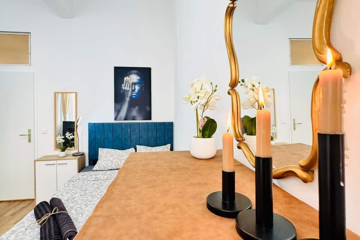 Een moderne slaapkamer met een grote spiegel, een bed met een blauwe hoofdbord, een nachtkastje met een lamp en een schilderij aan de muur.