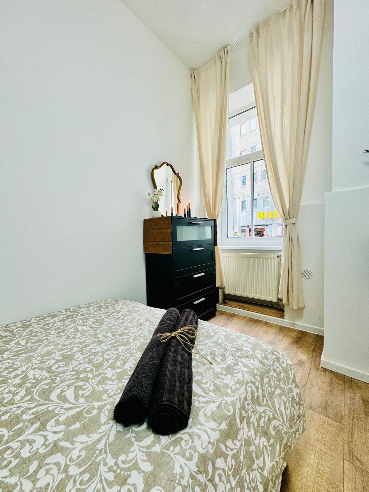 A bedroom with a bed, a dresser, a mirror, and a window with curtains.