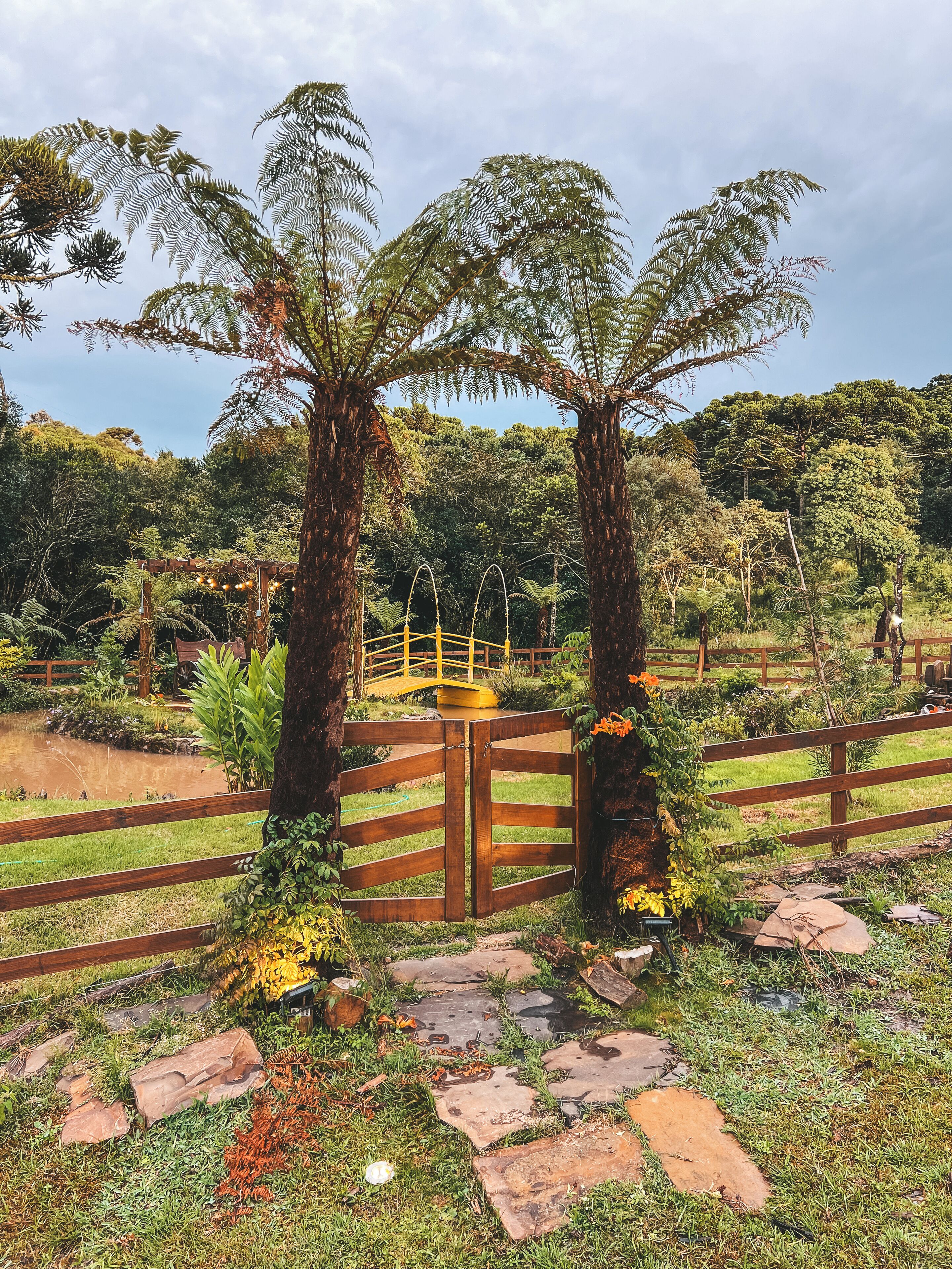 Um jardim com portão de madeira, palmeiras e uma ponte.