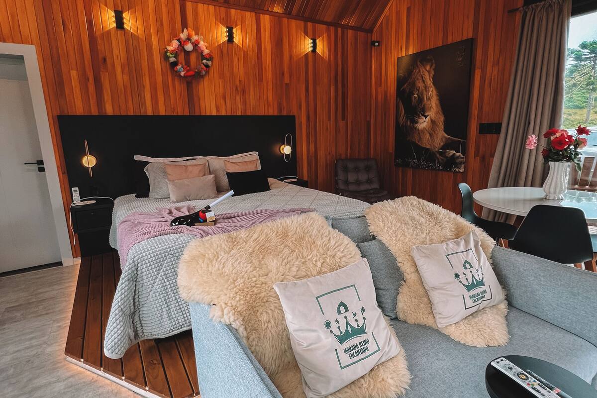 A modern bedroom with a wooden ceiling, a large bed with a headboard, a grey sofa with fluffy cushions, a small table with a remote control, and a painting of a lion on the wall.