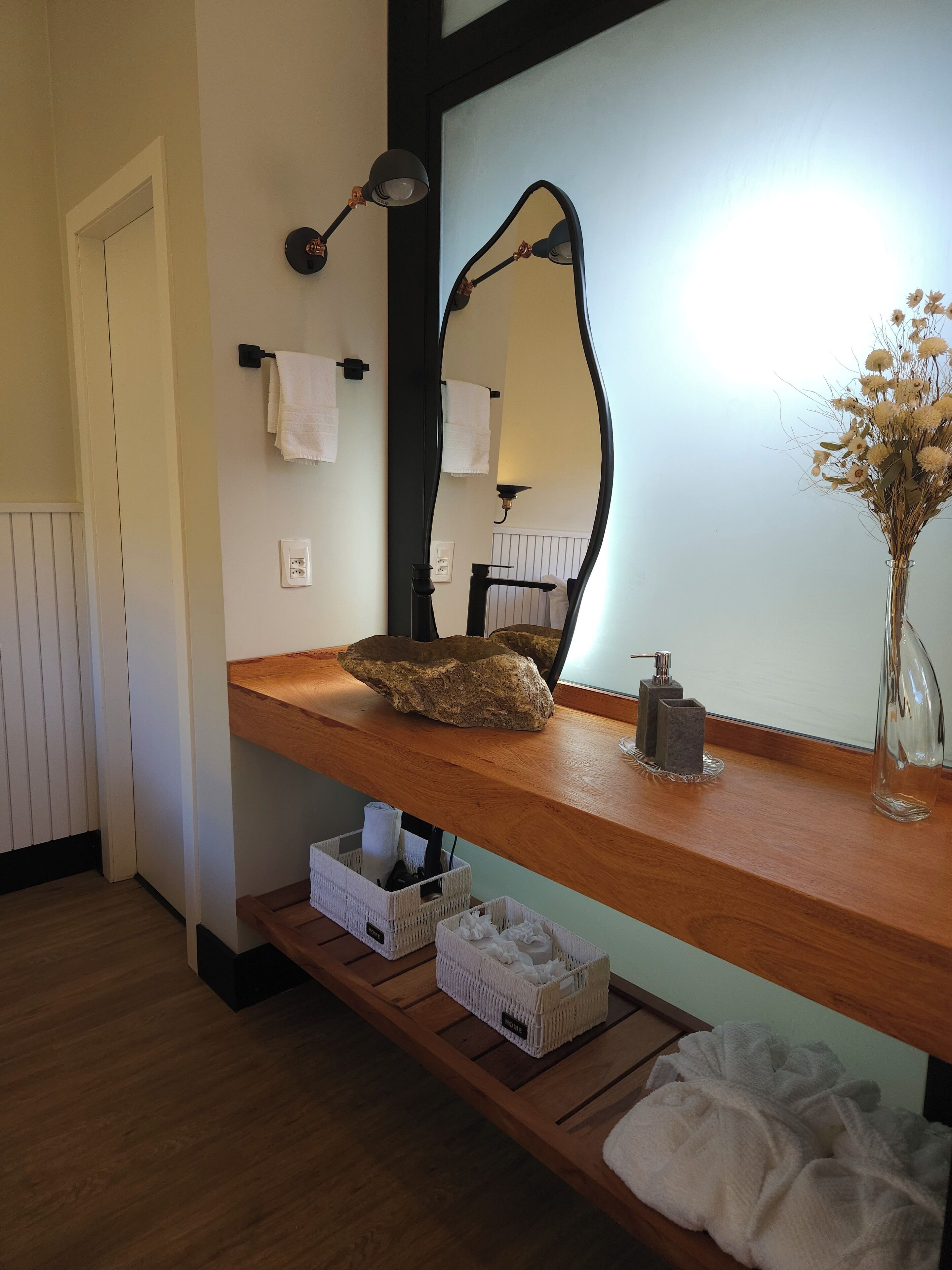 Un baño con tocador de madera, un espejo, un perchero, un jarrón con flores secas y una estantería con cestas.