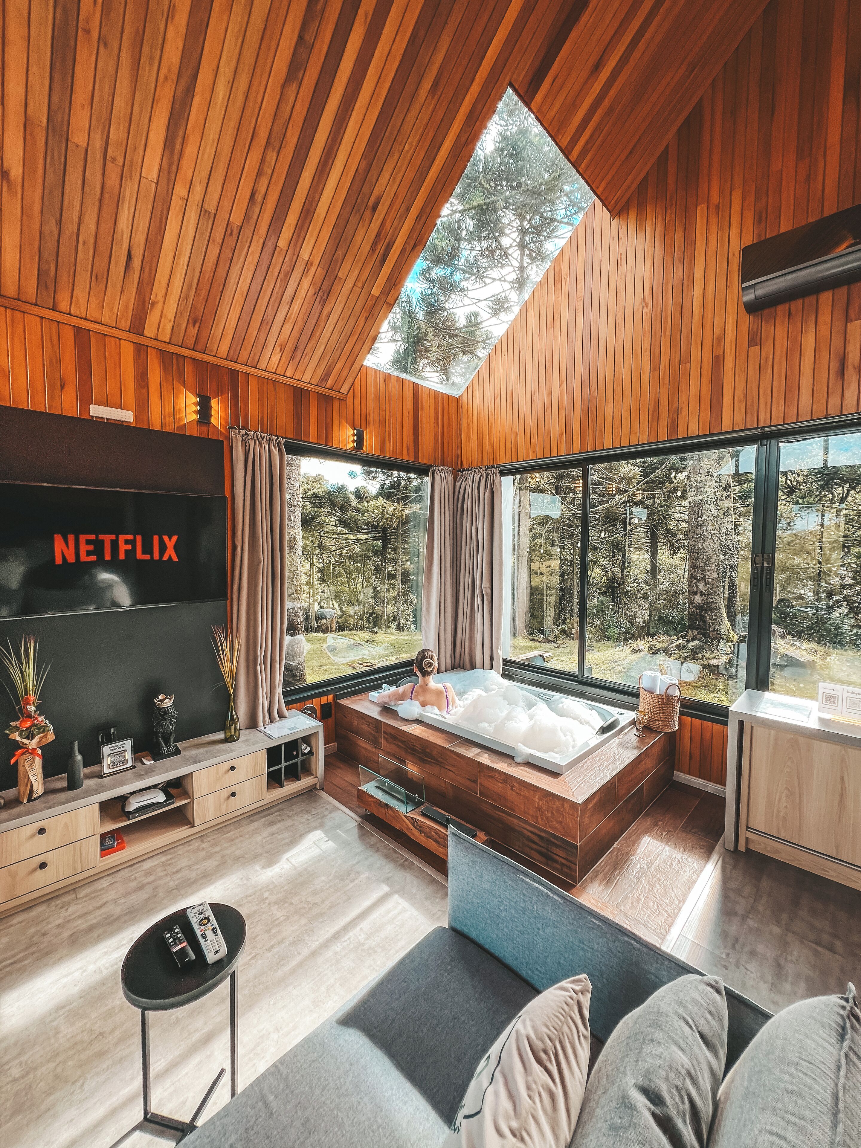 Une chambre moderne dotée d’une grande fenêtre, d’un plafond en bois et d’un téléviseur à écran plat sur lequel on peut lire le mot « NETFLIX ».