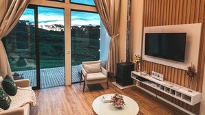 A modern living room with a large TV, comfortable seating, and a view of a natural landscape through large windows.