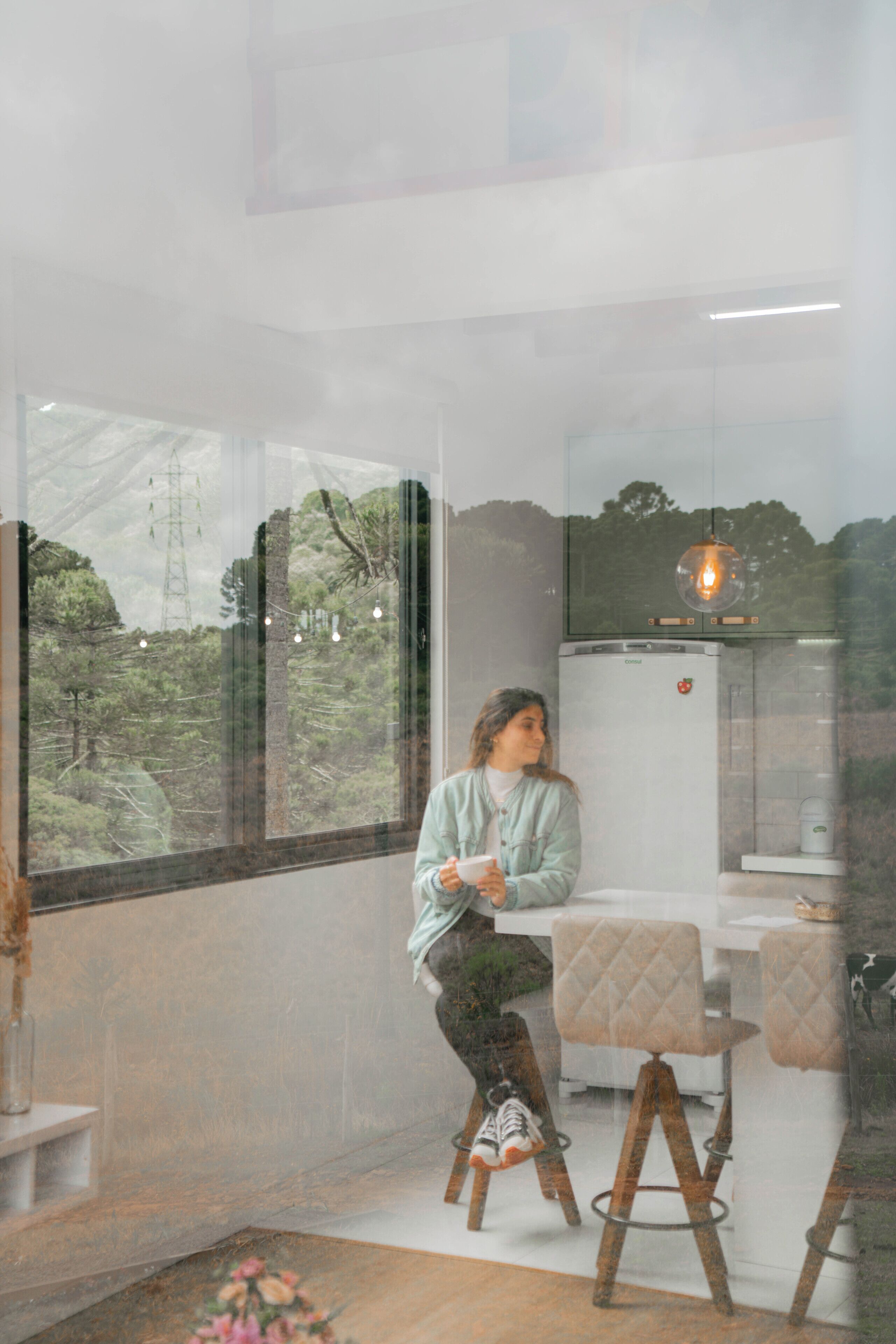 A person sitting on a stool in a room with a large window, a refrigerator, and a hanging light fixture.