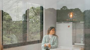 A person sitting on a stool in a room with a large window, a refrigerator, and a hanging light fixture.