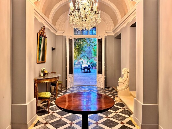 A grand hallway with a checkered floor, a round table, a chandelier, and a view of an outdoor garden.