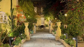A stone pathway leading to a courtyard with a table and chairs, surrounded by lush greenery and flowering plants. - Palazzo dei Fiori since 1883 (Balzan)
