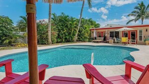 A swimming pool with red lounge chairs, a building with a red roof, and palm trees in the background.