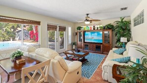 A spacious living room with a large TV, comfortable seating, and a view of a swimming pool.