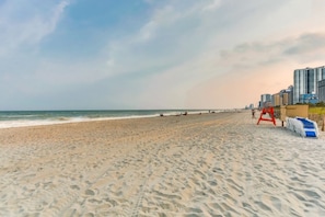 A beach with a lifeguard tower and umbrellas, a clear sky, and buildings in the background. - Palms Resort 501: 3BR Oceanfront Condo (Myrtle Beach)