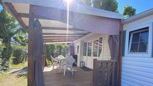 A covered outdoor deck with a table and chairs, a wooden fence, and a house with a window. - MOBILHOME 6 PERS CAMPING 3 Stars near the sea (Lunel)