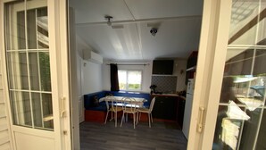 A compact living space with a dining area, kitchenette, and a window overlooking greenery. - MOBILHOME 6 PERS CAMPING 3 Stars near the sea (Lunel)