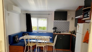 A compact kitchen with a dining area, a built-in oven, and a window with curtains. - MOBILHOME 6 PERS CAMPING 3 Stars near the sea (Lunel)