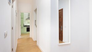 Un pasillo con suelo de madera y paredes blancas que conduce a una zona de cocina con armarios verdes y una puerta de madera.
