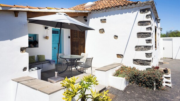A white building with a tiled roof, a wooden door, and a small garden with flowers.