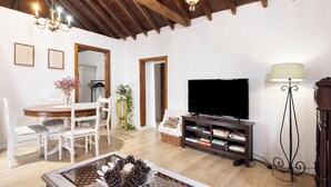 A living room with a wooden ceiling, a dining table with chairs, a TV stand with a flat-screen TV, a floor lamp, and a decorative table centerpiece.