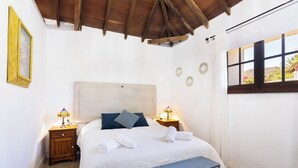 A bedroom with a wooden headboard, a bench, a bed with white bedding, two bedside tables with lamps, and a window with a view of greenery.