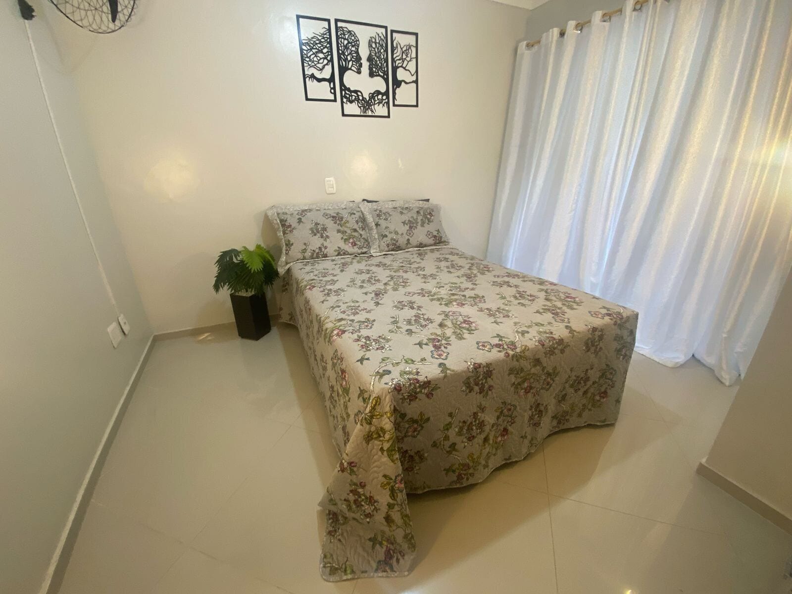 A bedroom with a floral bedspread, a potted plant, and a wall with three framed artworks.