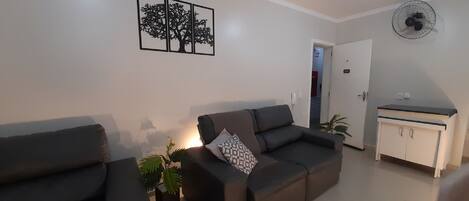A living room with a sofa, a small table with a plant, a wall-mounted fan, and a door leading to another room.