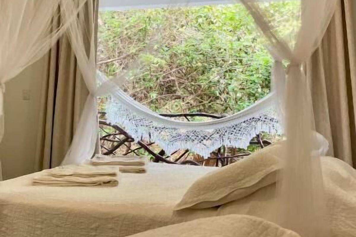 A room with a bed under a white canopy, a window with a view of greenery, and a small table with a lace tablecloth.