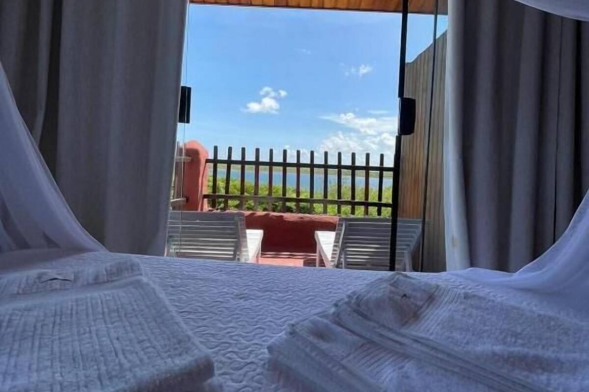 A bed with white bedding and sheer curtains, a wooden ceiling, and a view of a fenced area with a chair and a red object in the background.