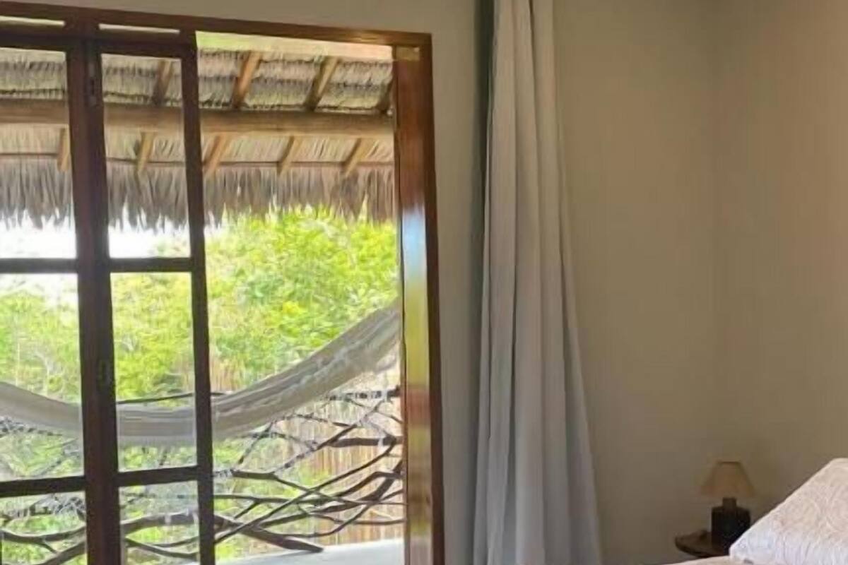 A bedroom with a bed, a nightstand, a lamp, and a view of a thatched structure outside.