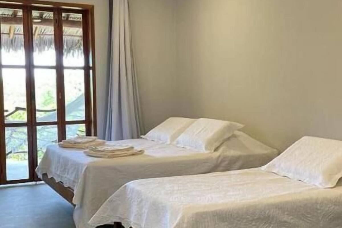 A room with two single beds, a large window with curtains, and a view of greenery outside.