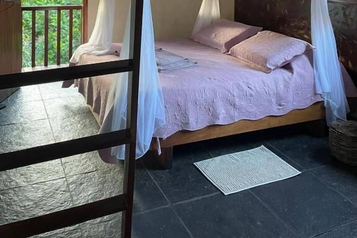 A room with a bed under a canopy, a balcony with a railing, and a view of greenery outside.