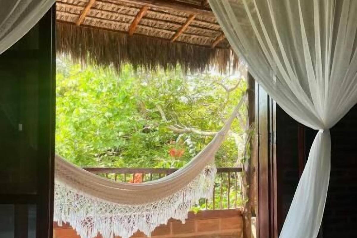 A room with a bed, a hammock, and a view of greenery through a window with white curtains.