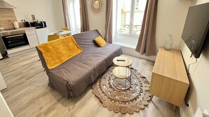 A modern bedroom with a sofa bed, a small table, a decorative rug, a TV stand, and a kitchen area in the background.