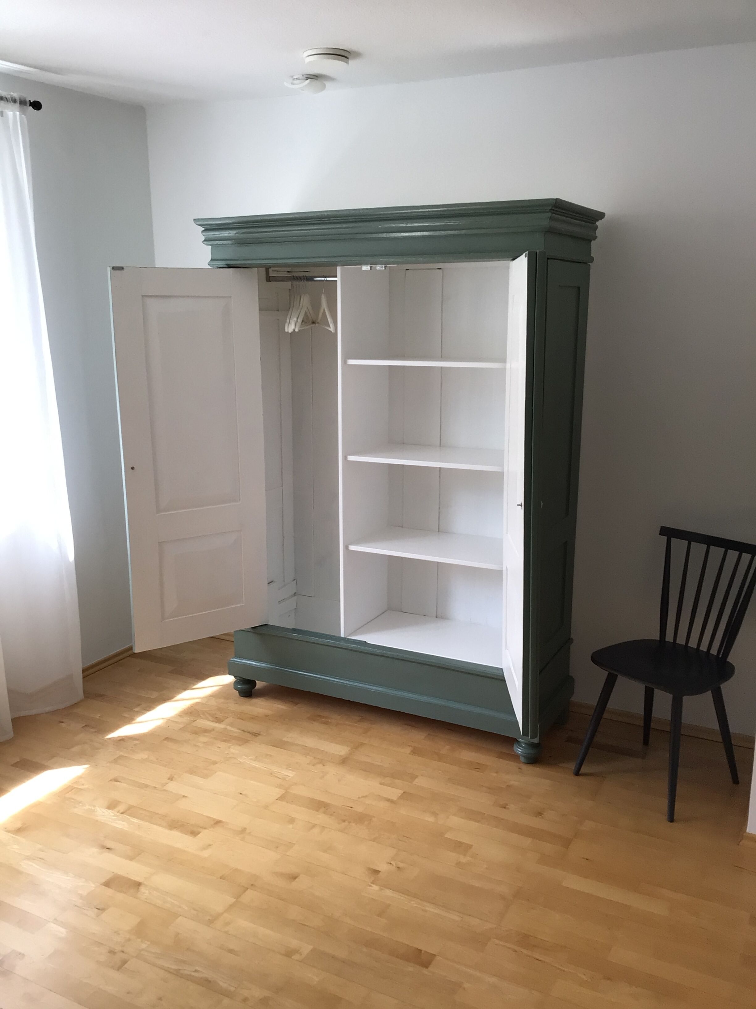 Ein Zimmer mit Holzboden, einer weißen Tür, einem grünen Kleiderschrank mit drei Regalen und einem schwarzen Stuhl.