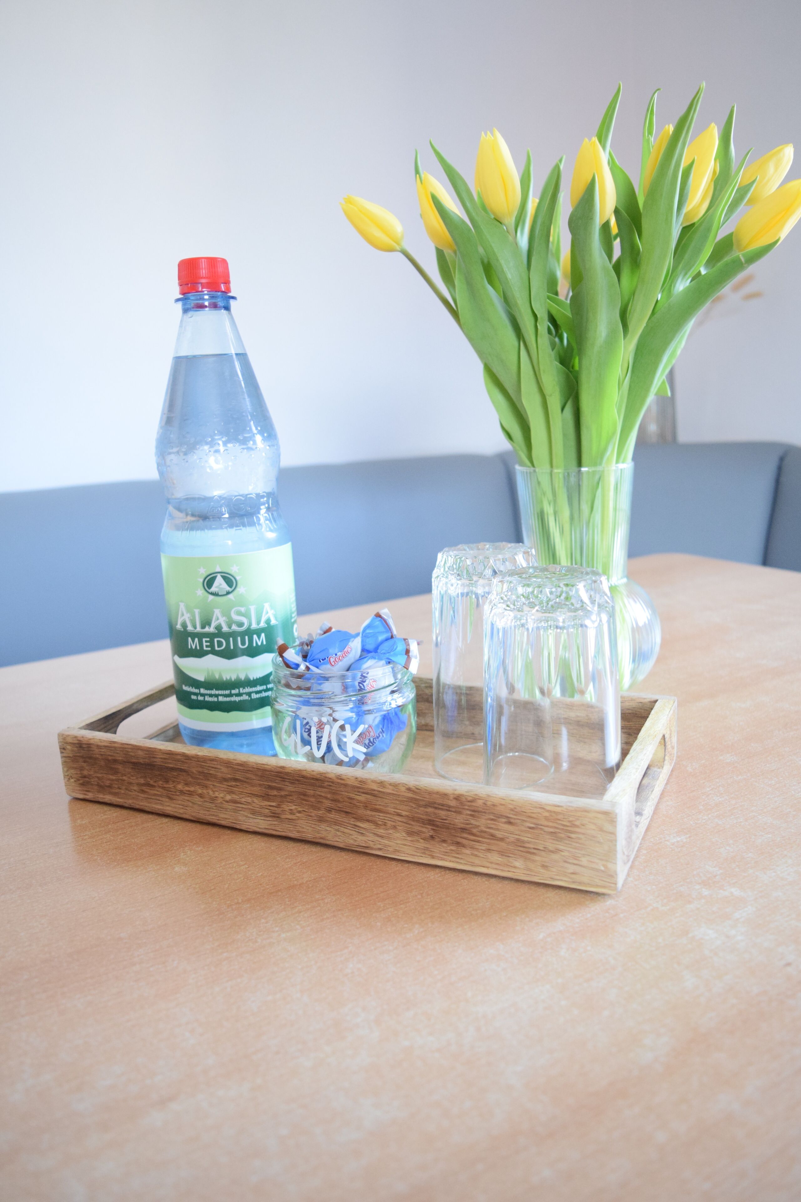 A wooden tray with a bottle of Alasia Medium water, a jar of candies, and a vase with yellow tulips on a table.