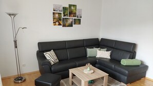 A modern living room with a black leather sofa, a wooden coffee table, and a black ottoman. There are four framed pictures on the wall.