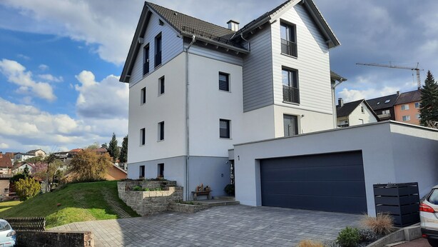 A modern two-story house with a garage, a stone retaining wall, and a paved driveway.