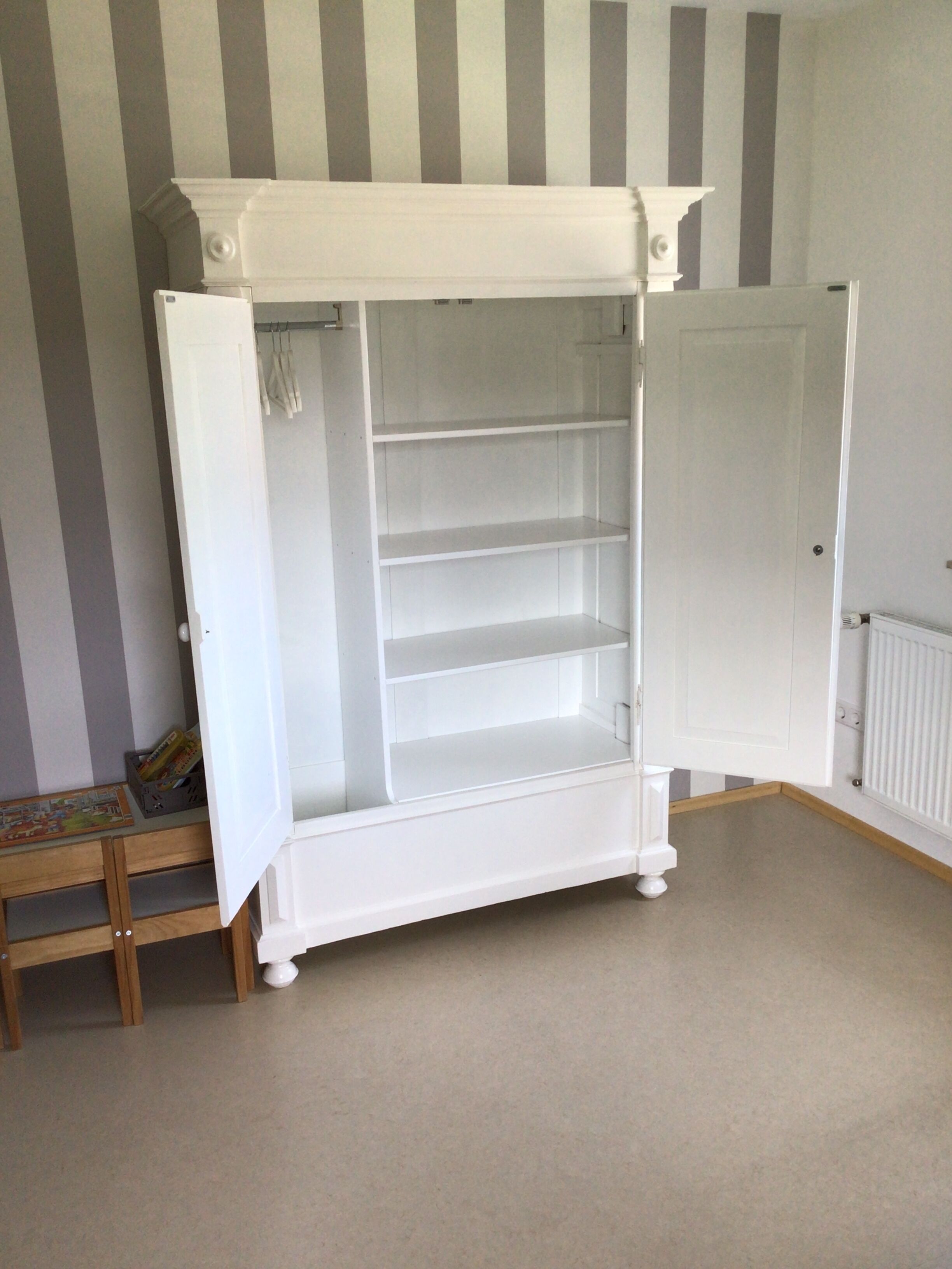 Een witte kast met meerdere planken en een deur, geplaatst in een kamer met gestreept behang en een radiator.