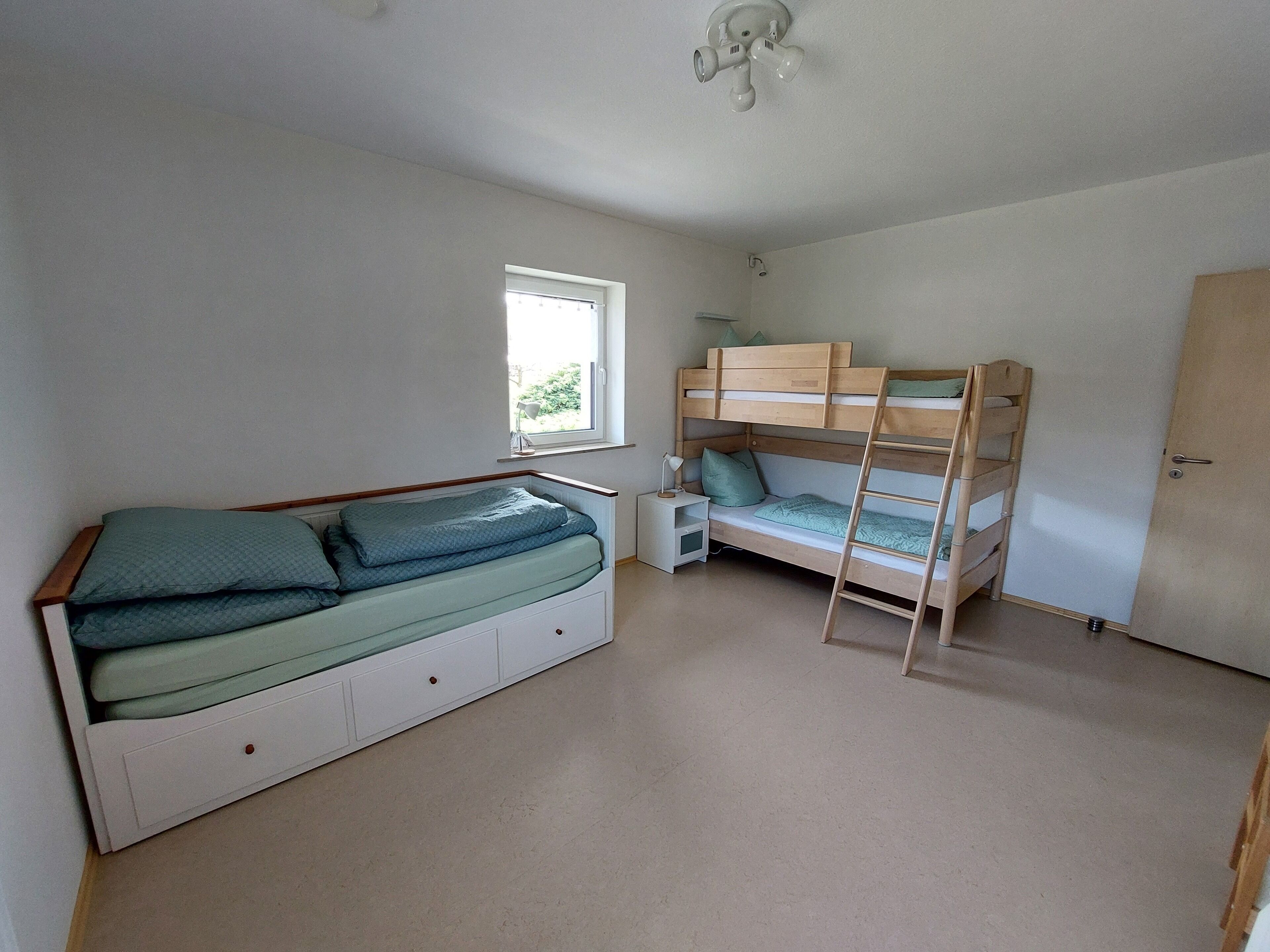 A bedroom with a bunk bed, a single bed, a bedside table with a lamp, and a window.