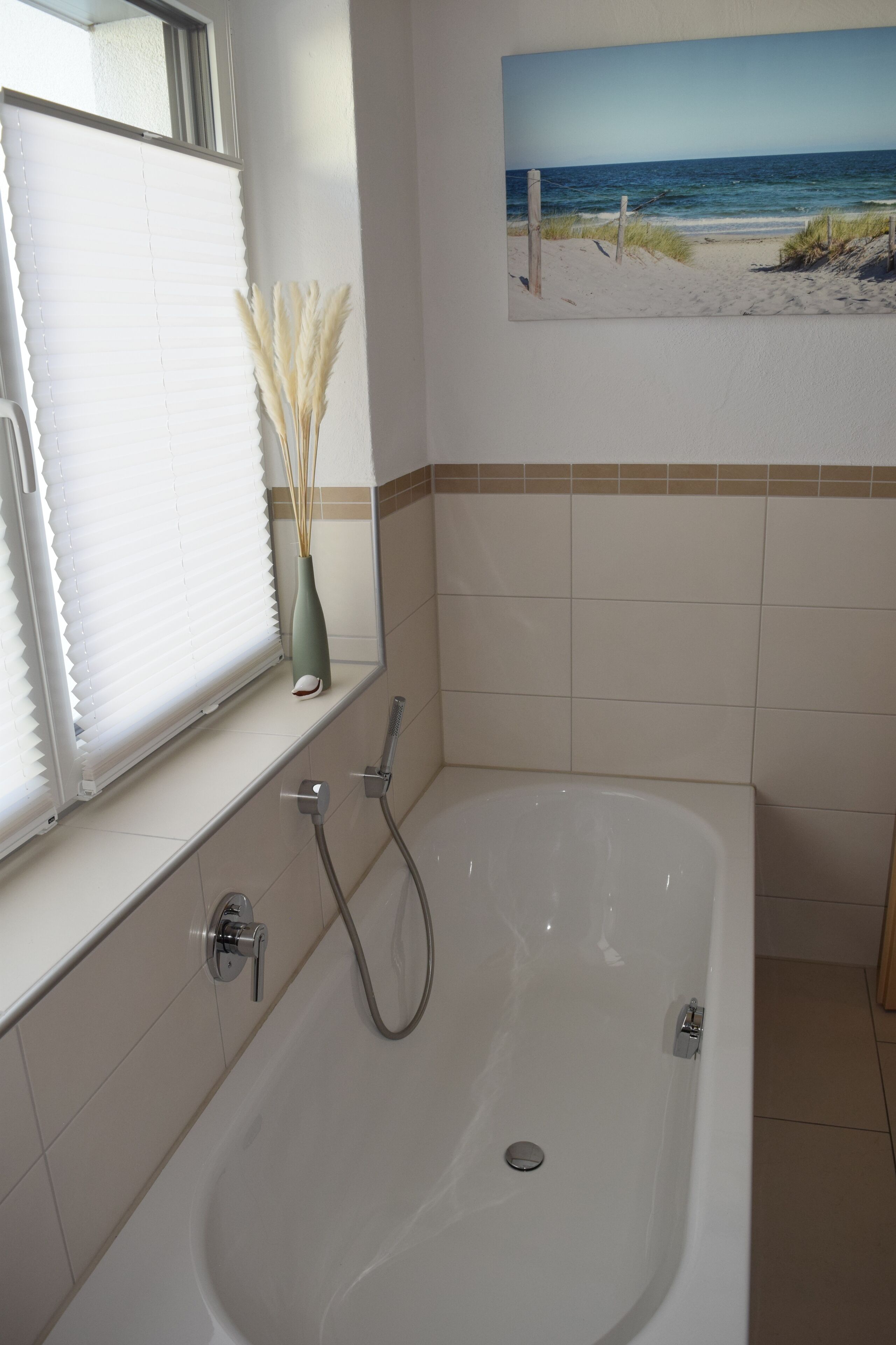 Une salle de bain avec une baignoire, une fenêtre avec des stores et un tableau sur le thème de la plage au mur.