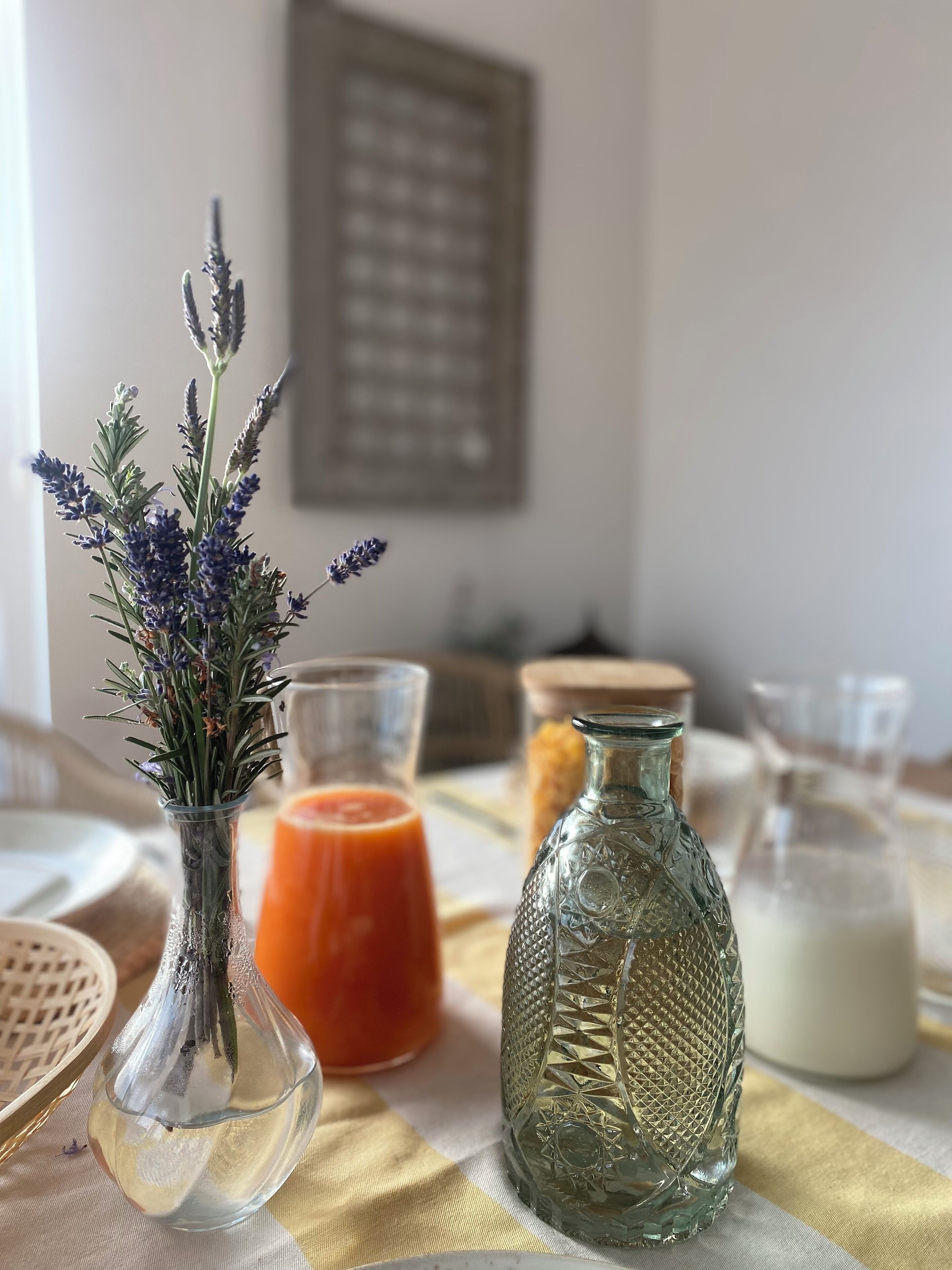 A table with a glass vase of lavender, a glass bottle of orange juice, and a glass bottle of milk.