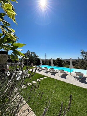 Una piscina con sillas de descanso y sombrillas, rodeada de un césped bien cuidado y plantas de lavanda.