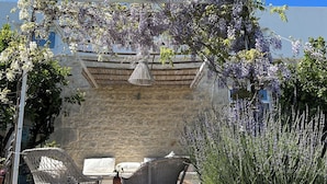 A stone wall with a pergola covered in purple flowers, wicker furniture, and a stone building in the background.
