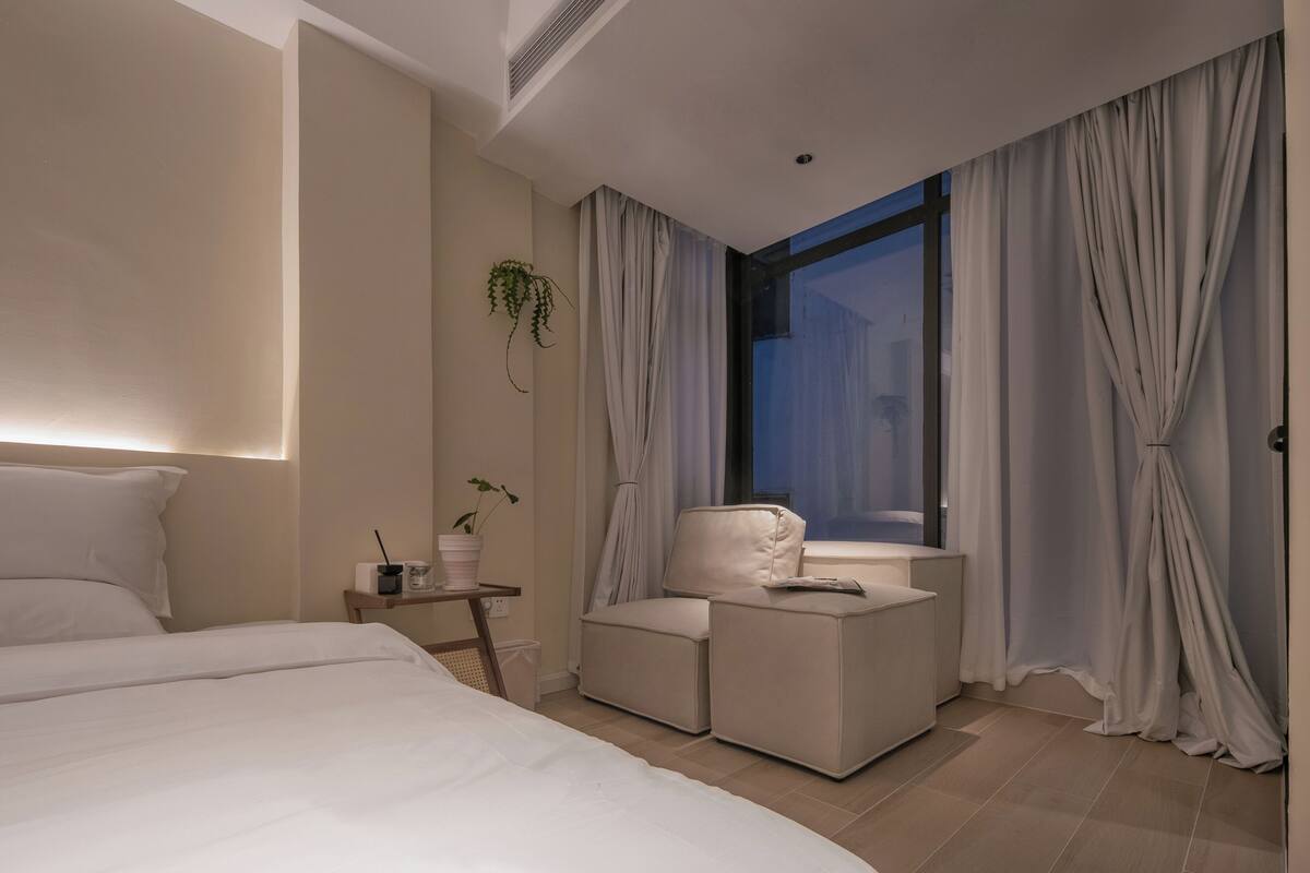 A modern hotel room with a large bed, a small seating area, a window with curtains, and a plant on the wall.
