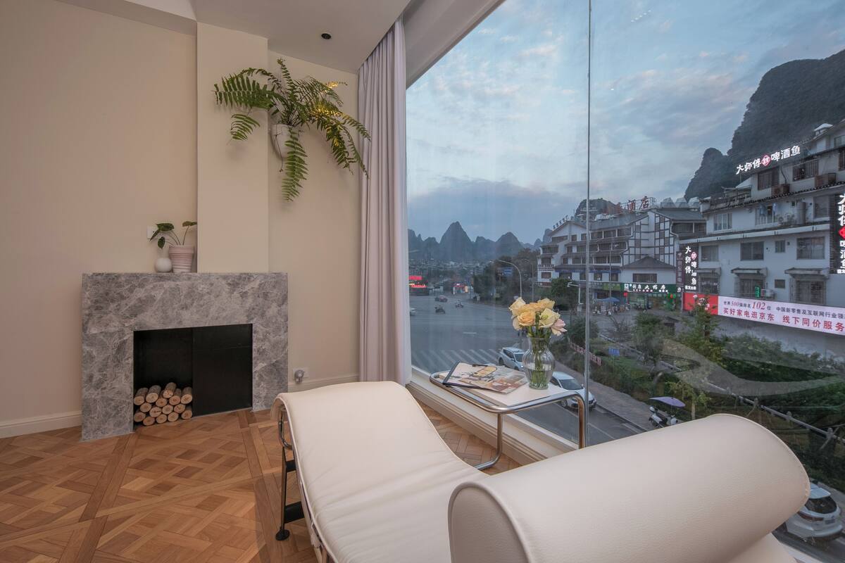 A modern hotel room with a large window, a fireplace, a massage chair, and a view of a cityscape with mountains.